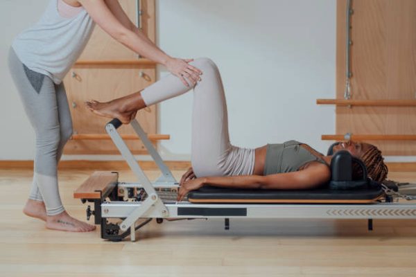 Pretty woman in sportswear doing pilates exercise on reformer with trainer assisting.